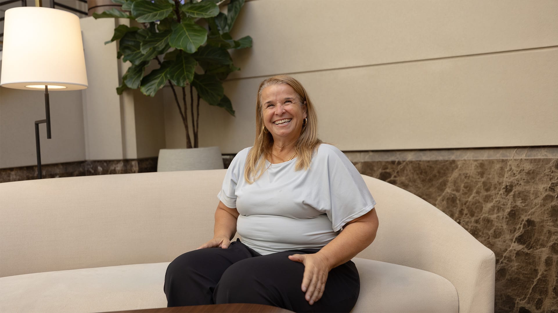 Yodi Richeson seated on a curved sofa, smiling, in a bright plant-filled room.