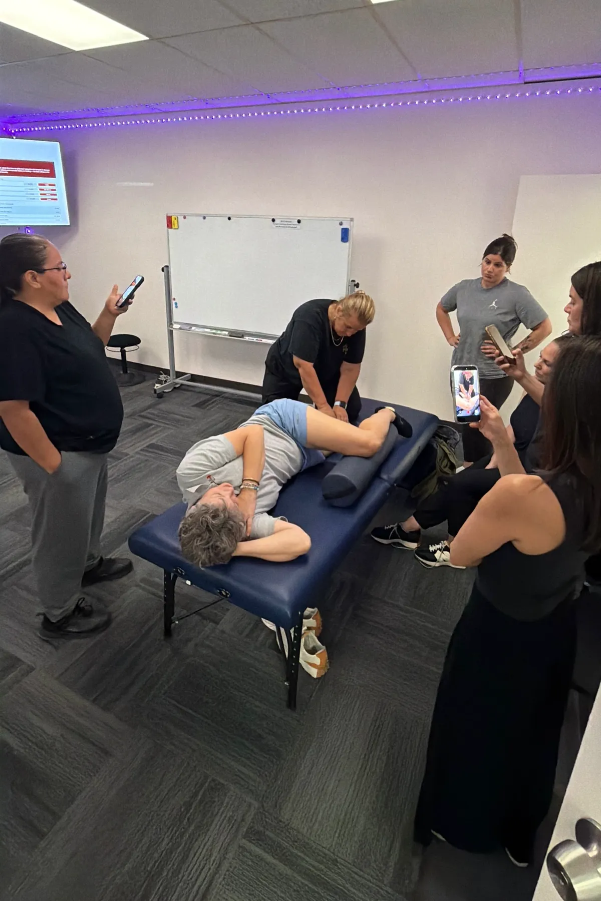 Yodi Richeson demonstrating a hip technique on a client while a group of practitioners watches and films
