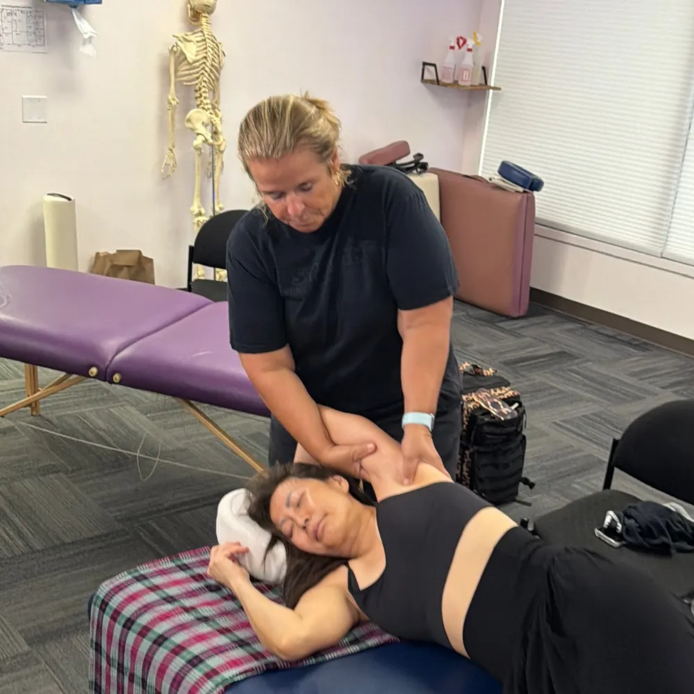 Yodi Richeson working on a client's shoulder on a treatment table, an anatomical skeleton model behind her